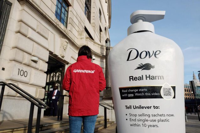 A giant Dove pump bottle stands in front of a white London building. A Greenpeace activist in a red jacket stands next to it, and it towers over her at around twice her height. The bottle reads "Dove Real Harm" "Real Change starts with YOU. Watch this video" with a QR code. It also reads "Tell Unilever to: Stop selling sachets now. End single use plastic within 10 years"