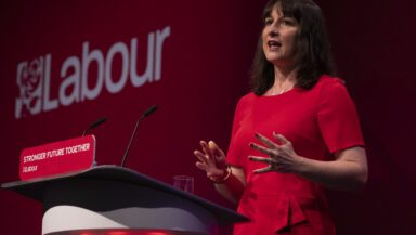 A woman with dark hair in a red dress stands giving a speech in front of a large "Labour" sign, at a lectern reading Stronger Future Together