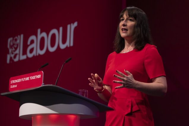 A woman with dark hair in a red dress stands giving a speech in front of a large "Labour" sign, at a lectern reading Stronger Future Together