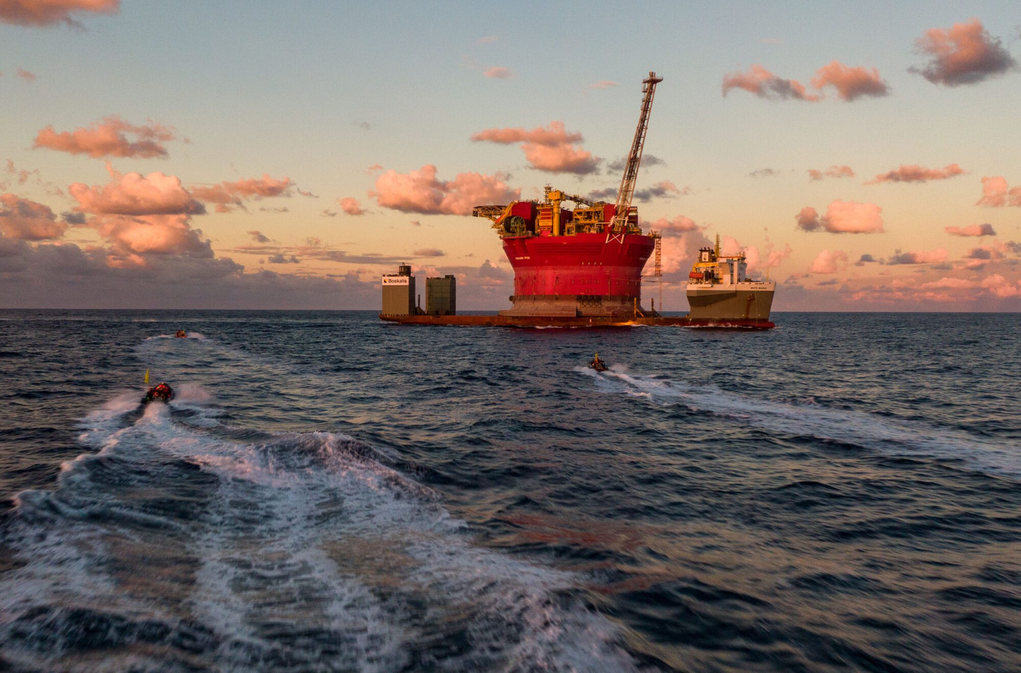 Three tiny rhib boats with white water trailing behind them approach a giant red oil platform with a large crane rising up to double its height, and two additional structures on each side. The platform are sat on the horizon of a still seascape at sunset, with pink clouds framing the structures.