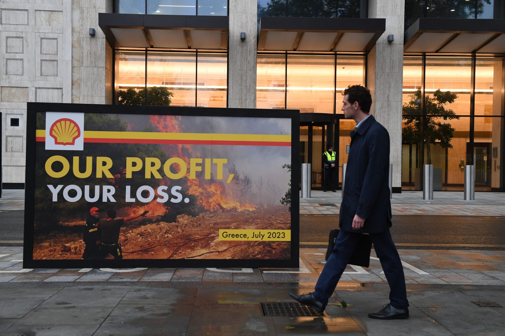 A man in a suit walks past a large billboard with a Shell logo in front of a fire scene, reading "Our profit, Your loss", in front of the entrance to a large office building with a double-height reception glowing with lights.