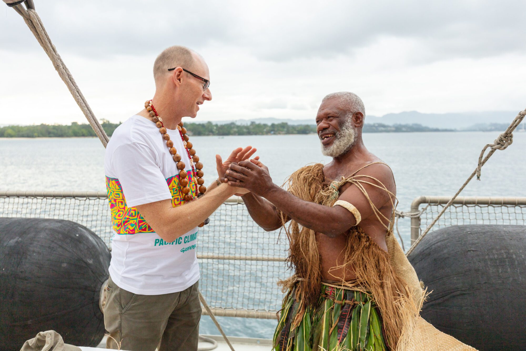 A white man in a white t-shirt stands facing a man of colour with white beard wearing a garment made of straw. They are smiling and clasping each others hands.