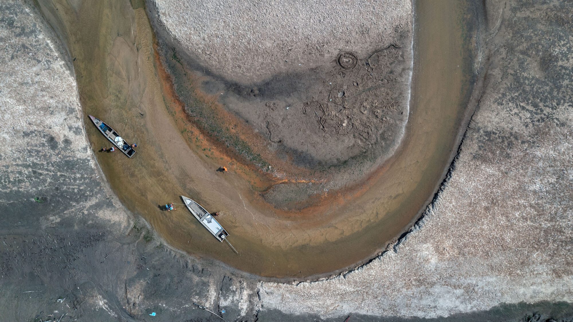 A dried out riverbed U-bend in brown with no vegetation or any signs of life around it holds two boats which have been beached in the sand of the riverbed