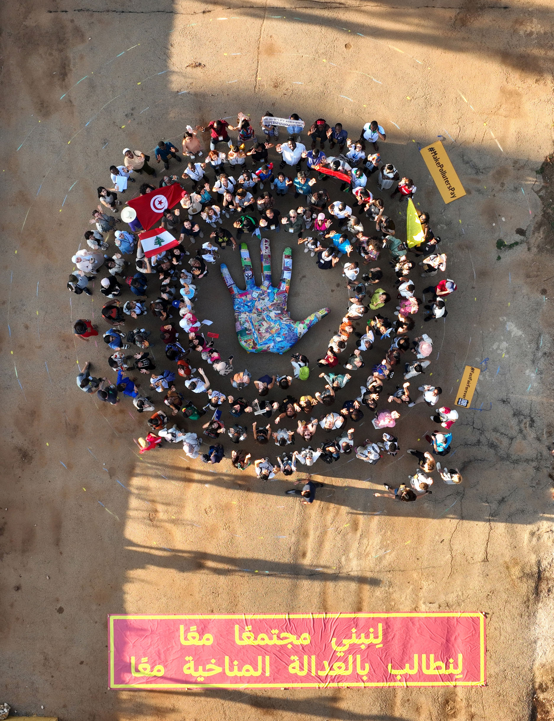 A bird's eye view of a crowd of people arranged in a circle around a blue hand. Underneath is a red banner with an Arabic text printed in yellow.