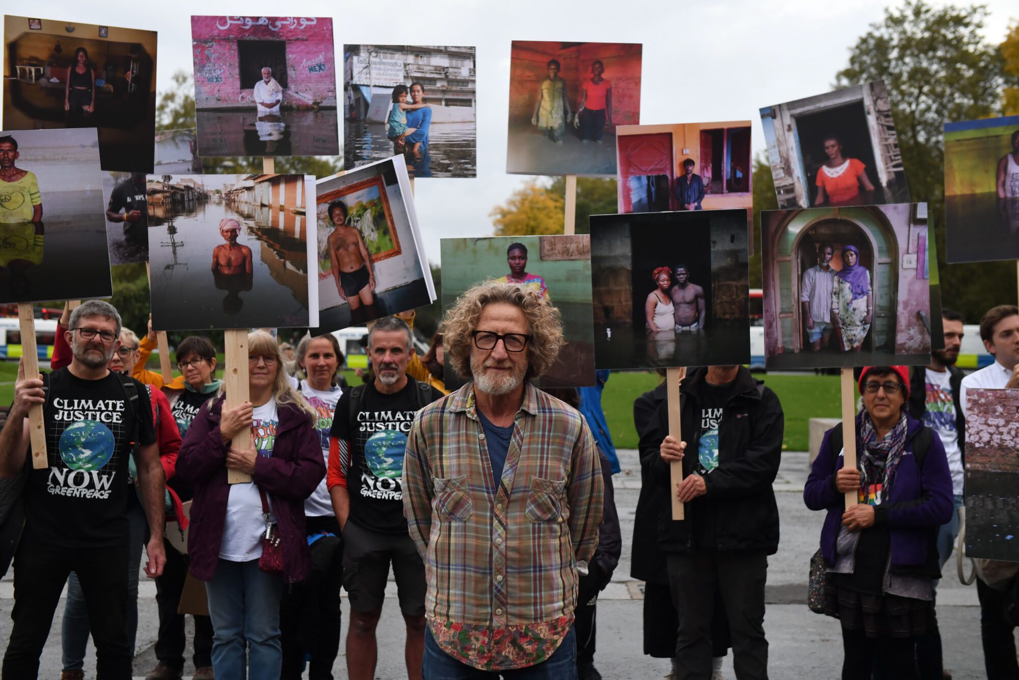 A crowd of people stand holding photographs high in the air, many wearing the same black "climate justice NOW!" T shirt, standing behind a smiling man with a beard and longish curly light brown hair.