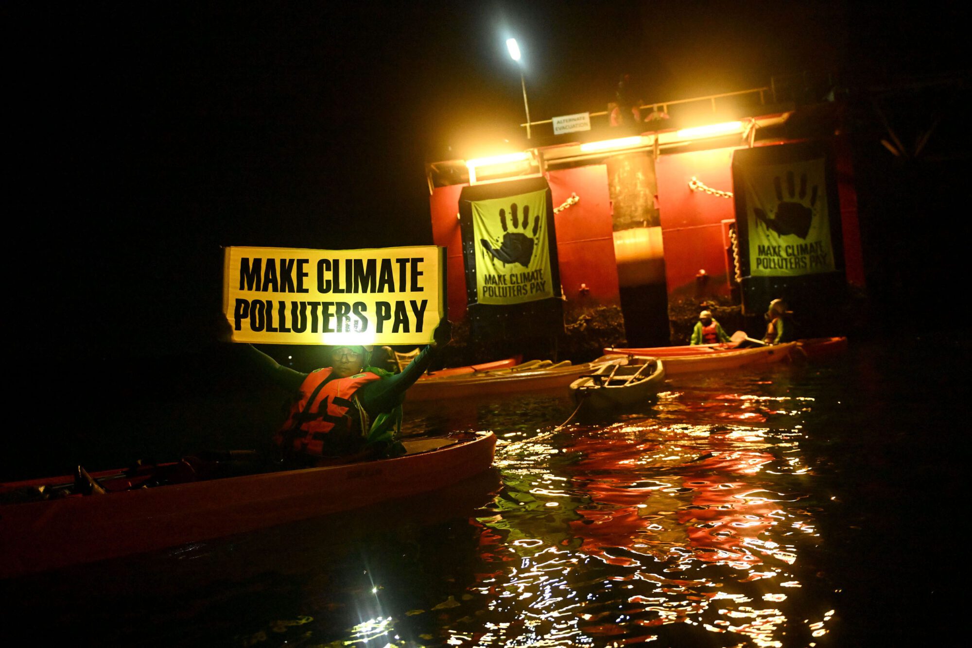 A night-time scene on water lit by a red vessel's bright yellow lighting. On the water are around six kayaks and an activist holding a yellow banner uplit to read the words "Make climate polluters pay". On the red structure under the lights are hung posters in yellow, both with black handprints and "make climate polluters pay" text underneath.
