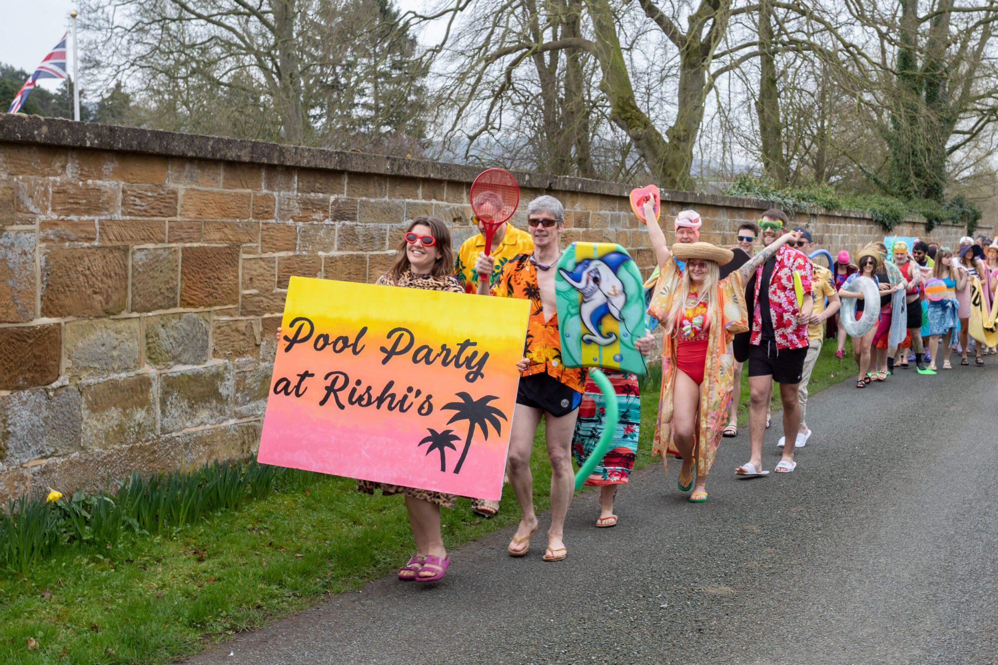 Against a brown wall, grey sky with leafless trees, a row of people in very colourful costumes, including flip flops and goggles and pool toys. A sunset-coloured banner reads in tropical-themed writing "Pool Party at Rishi's" with two palm tree motifs