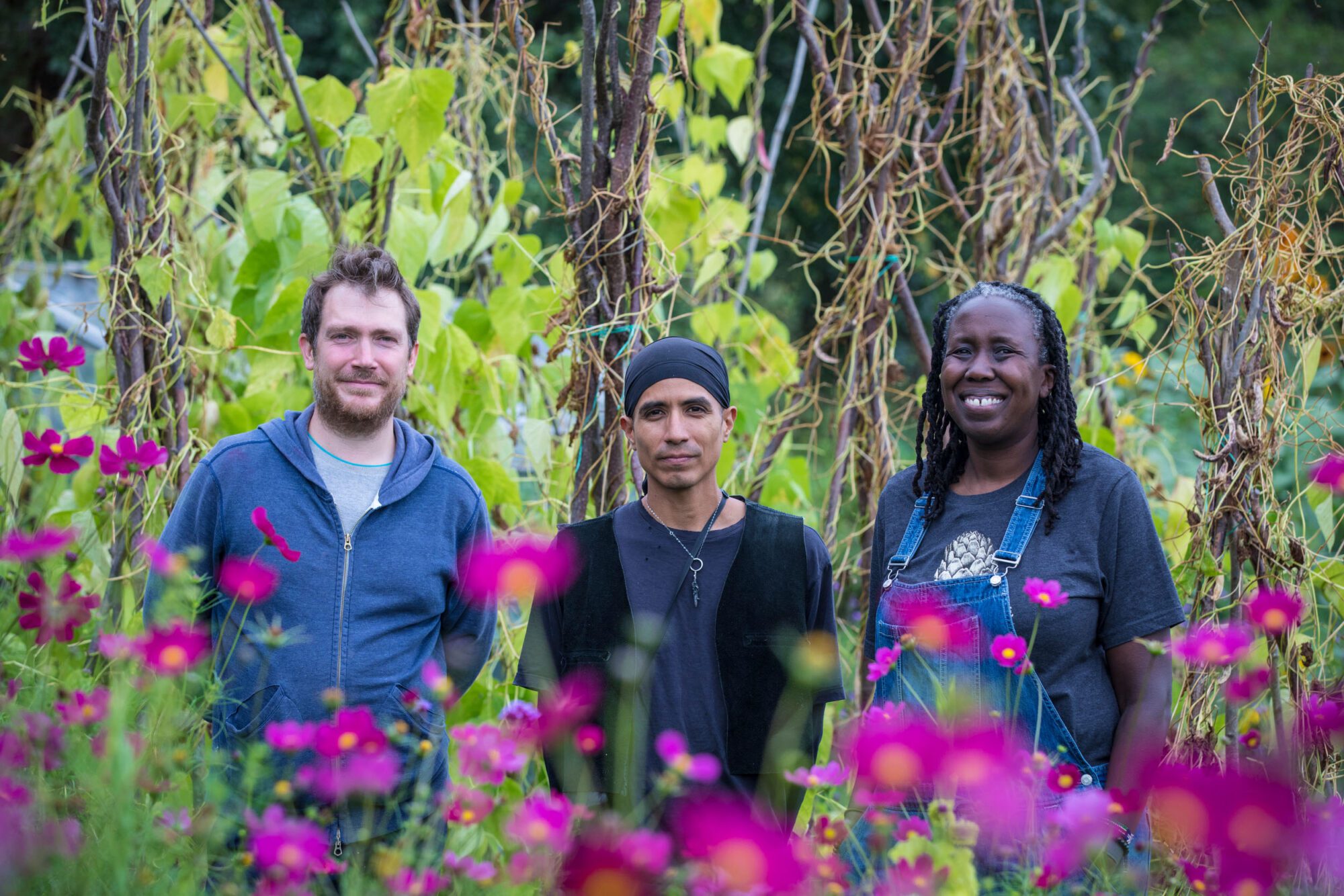 Three people – a white man, a brown man and a Black woman smiling – stand against a lush green plant background and bright pink flowers in the foreground.