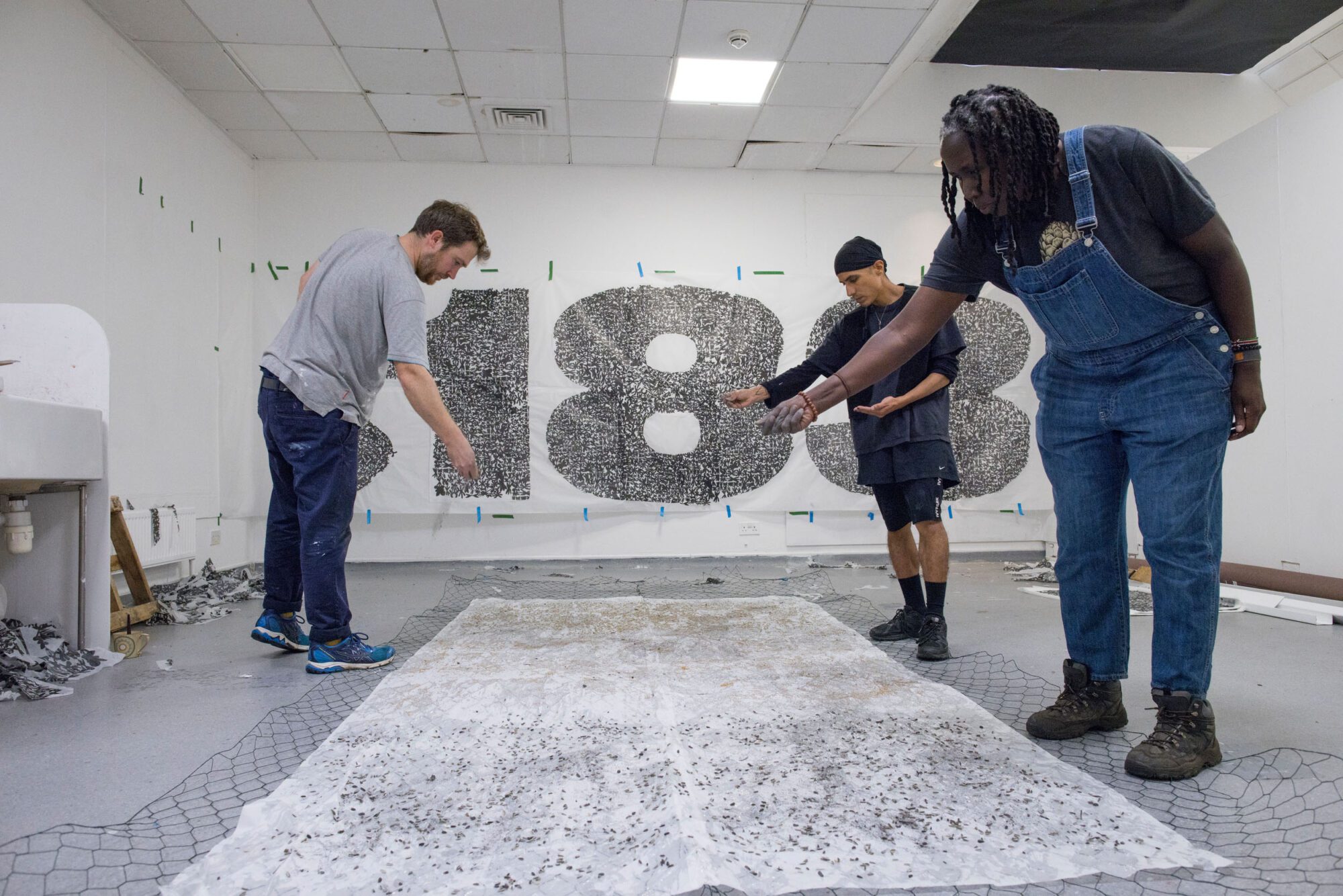 IN a white room three people stand over a white sheet scattering seeds and dust. In the background hands a white banner with giant numbers 1 and 8 printed with a grey pattern