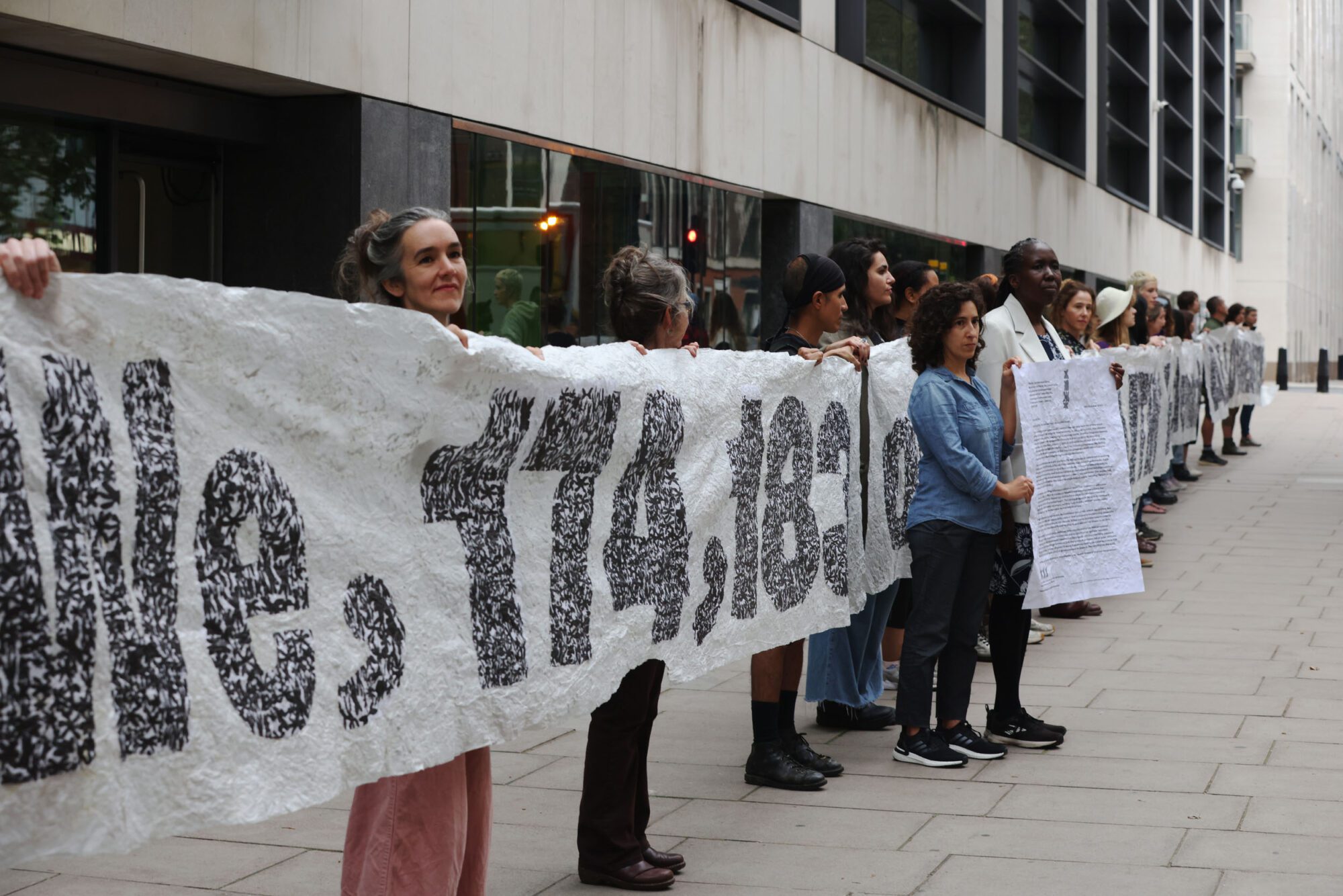 A long line of people stand in front of a big building, holding a white banner with grey lettering banner reading "We, 174,183..." becoming illegible thanks to its length and people standing in front holding a large piece of white paper