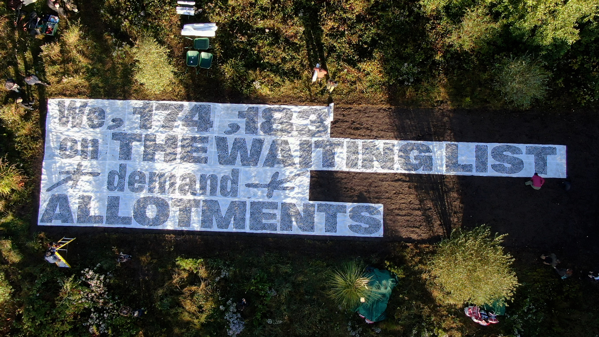 A birds-eye view of a very large and long white banner is split into four lines to be laid on a piece of brown land, recently ploughed and surrounded by green shrubs, reading "we, 174,138 on THE WAITING LIST demand ALLOTMENTS". There are a couple of people and ladders around showing the scale