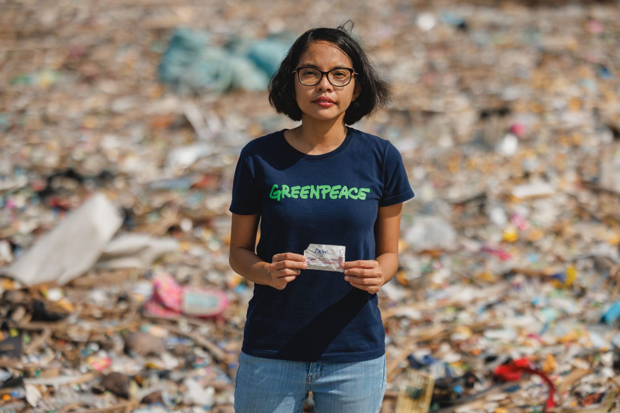 A woman in a Greenpeace t-shirt stands holding a small piece of plastic reading Dove against a background filled entirely with plastic waste
