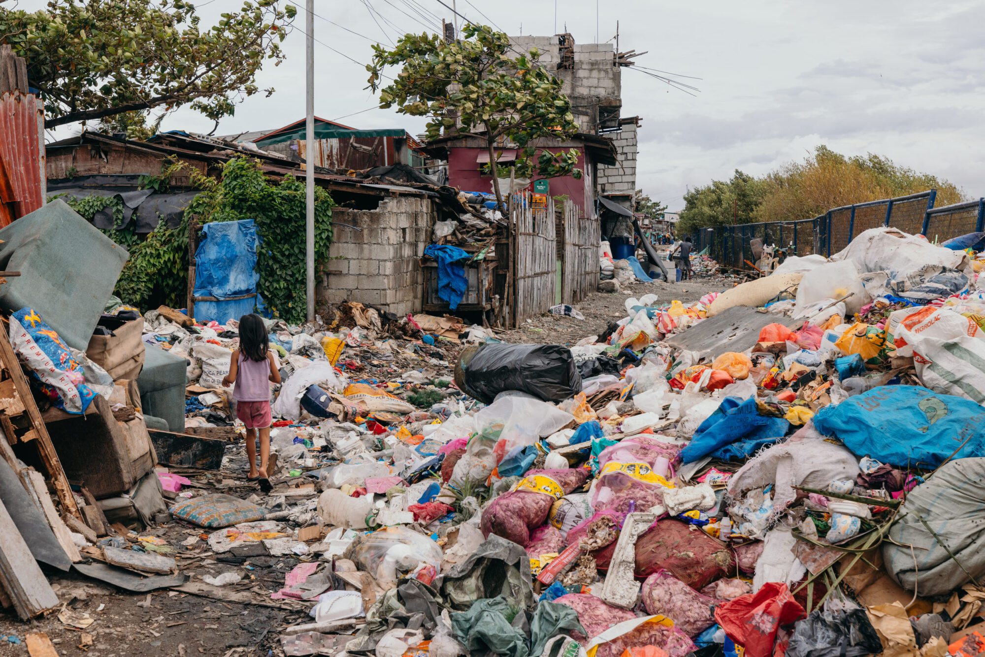 A village street is completely filled with plastic rubbish and bags of all colours and sizes, which is piled high on the right side of the road. A small girl is walking over rubbish towards the shack-link buildings just beyond.