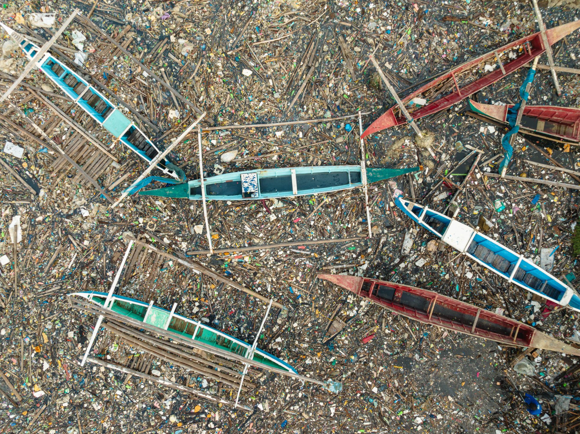 A birdseye view of thin boats in green blue and red; the ground around them is littered with colourful small bits of plastic