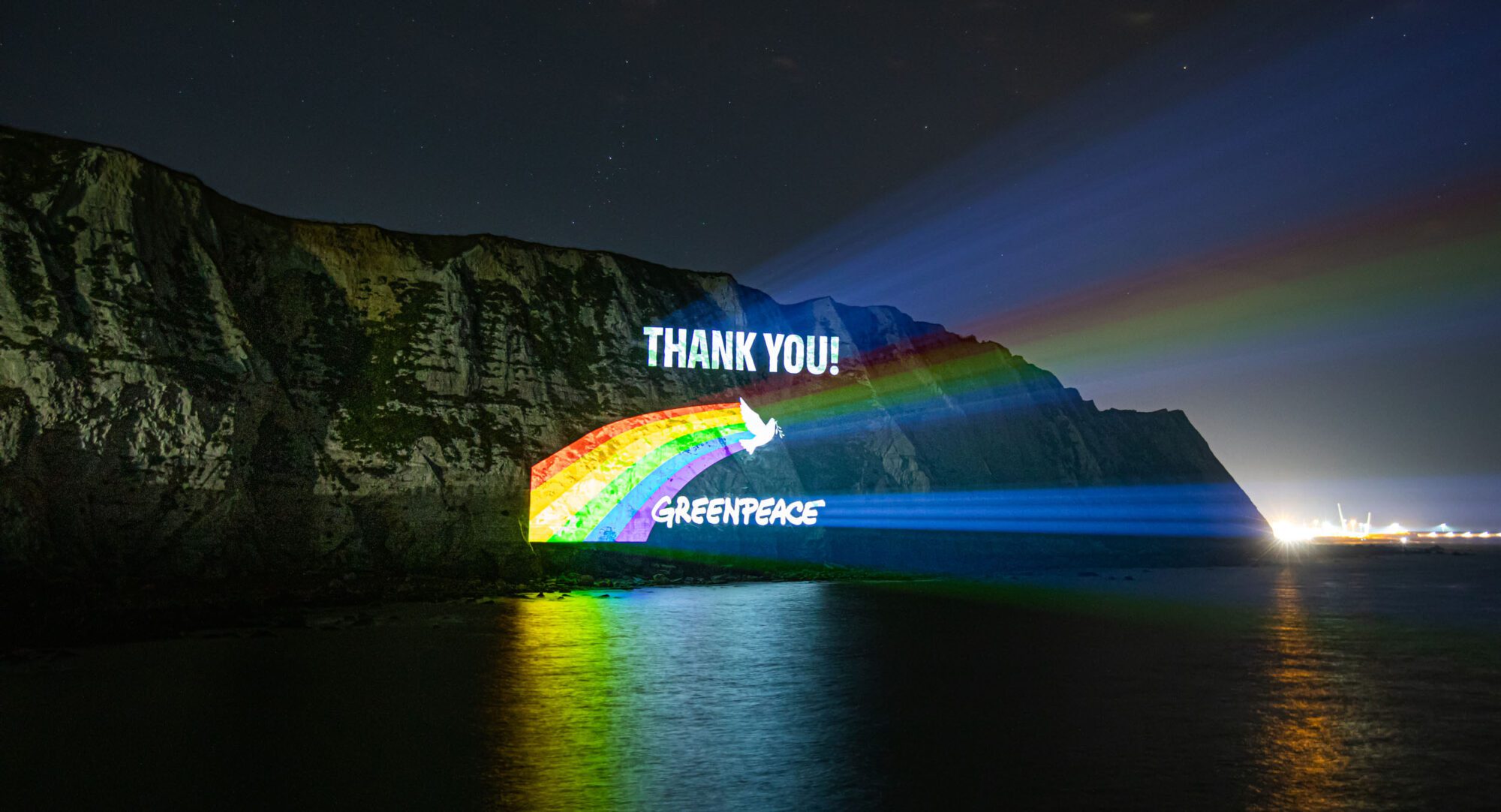 An image of a cliff at night with a projection reading "Thank you" with a rainbow streaming from behind a white bird. and the word Greenpeace
