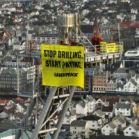 On a background of dense greyscale cityscape filling the entire frame, protestors are holding a yellow banner reading ‘Stop drilling. Start Paying. Greenpeace' high above on a metal frame