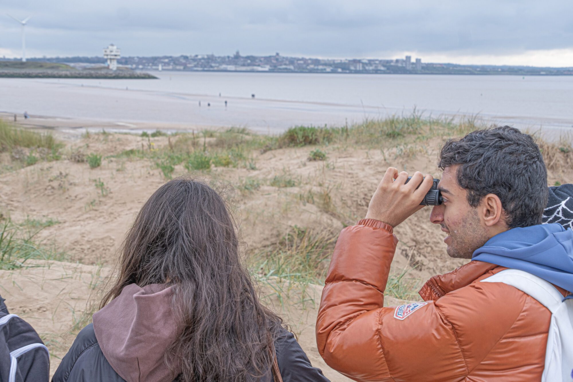 Two people walk among sand dunes, observing at a distant port across the water. One looks through binoculars.