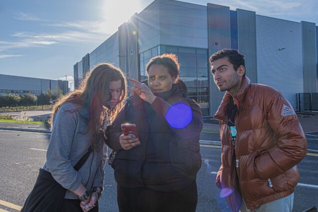 Three people of colour lean over a phone screen on a wintery light industrial estate. One points at something off camera.