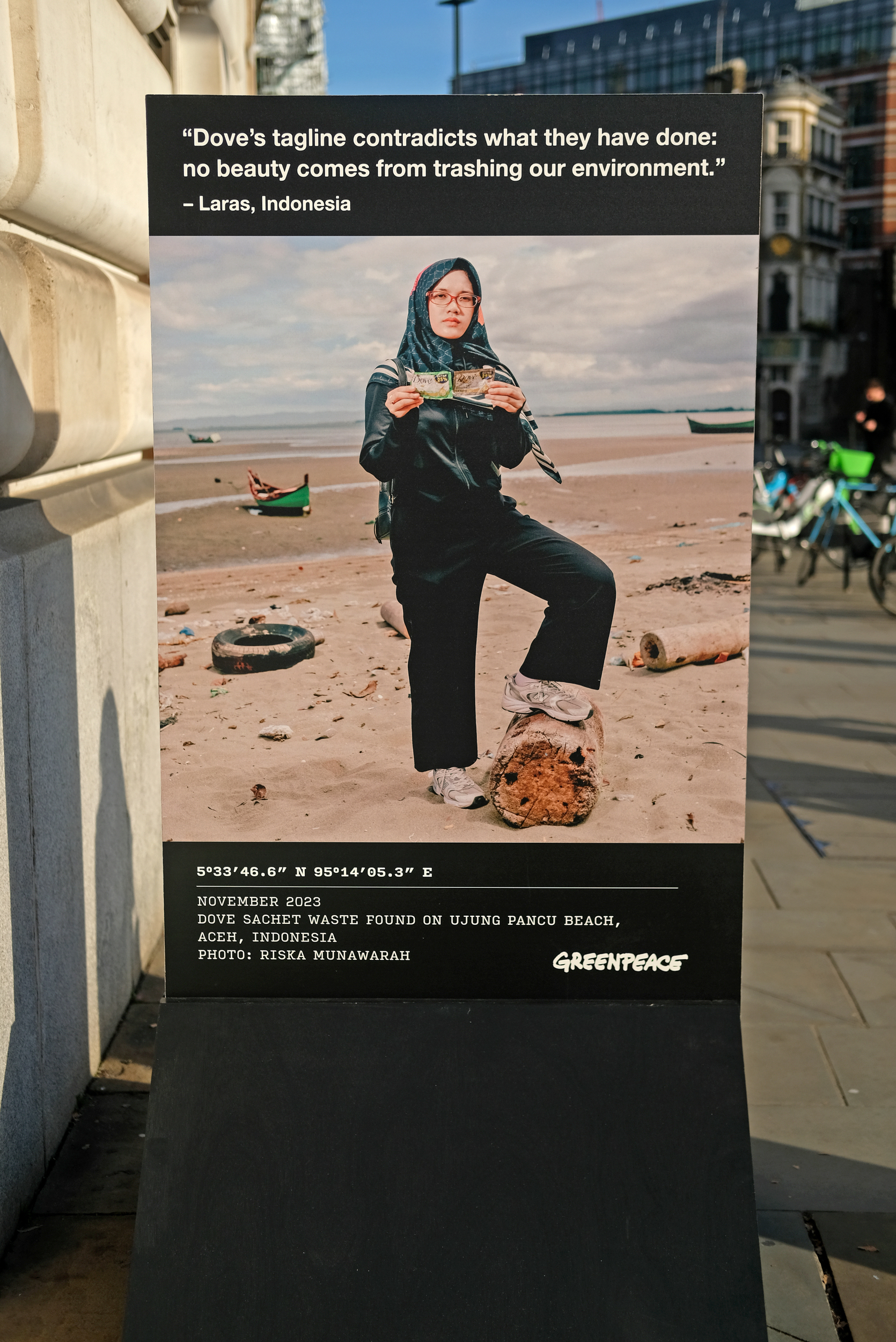 A standup sign in the street showing a woman on a beach holding up a littered Dove sachet. Quote reads "Dove's tagline contradicts what they have done: no beauty comes from trashing our environment."