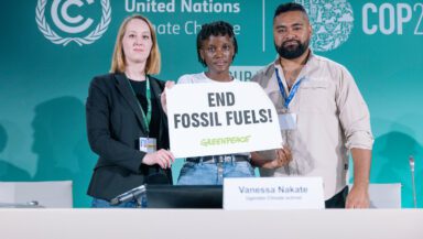 3 people stand on a platform with text behind and in front of the desk reading United Nations COP28UE with a small banne reading End Fossil Fuels! Greenpeace. There is a name tag in front of the woman holding the banner reading Vanessa Nakate