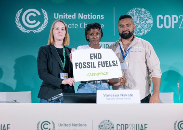 3 people stand on a platform with text behind and in front of the desk reading United Nations COP28UE with a small banne reading End Fossil Fuels! Greenpeace. There is a name tag in front of the woman holding the banner reading Vanessa Nakate