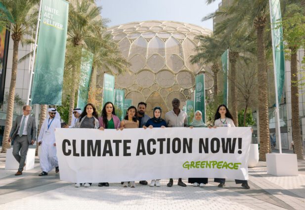 A group of young activists hold a large banner that says 'climate action now'. They're in a bright plaza lined with palm trees with an ornate conference building in the background. A group of men in traditional emirati dress or western business suits walk past.