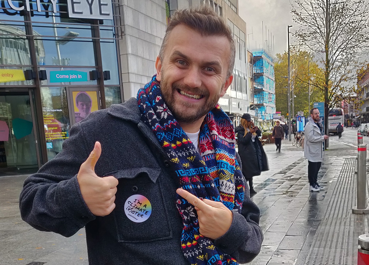 A friendly looking guy pointing at a sticker on his coat that says I'm a climate voter.