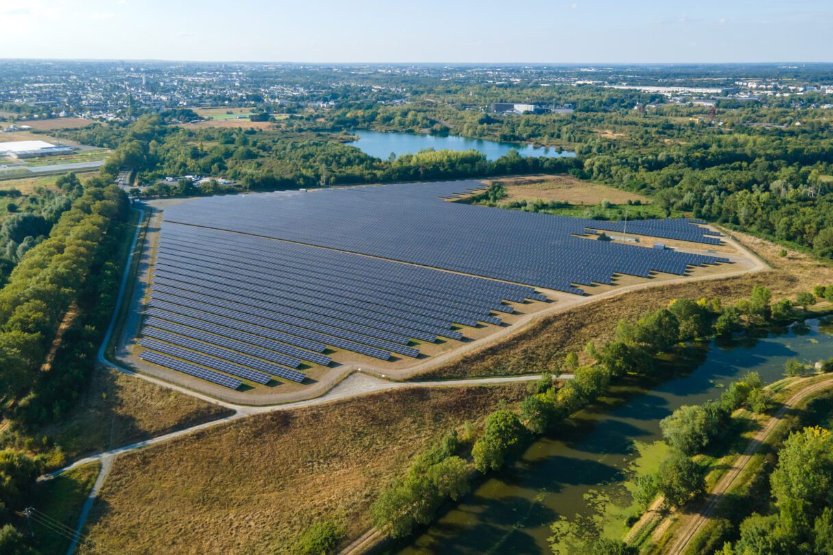 An aerial shot of a large field of solar panels, surrounded by trees and a small lake, with some houses and other neighbourhood buildings dotted across the horizon in the distance