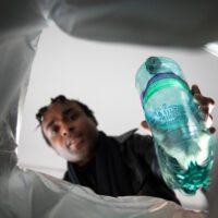 View from inside a bin, a person putting a green plastic bottle into it, framed by the plastic bag lining the bin.