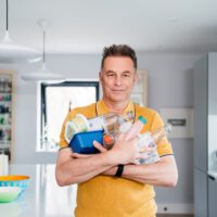 A man in a bright yellow T-Shirt stands in a kitchen with his arms full of plastic contain