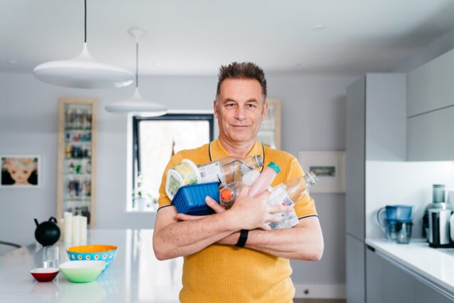 A man in a bright yellow T-Shirt stands in a kitchen with his arms full of plastic contain