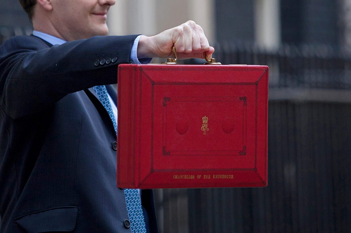 Closeup of the iconic 'red box' briefcase representing a UK budget being held out by a man in a suit.