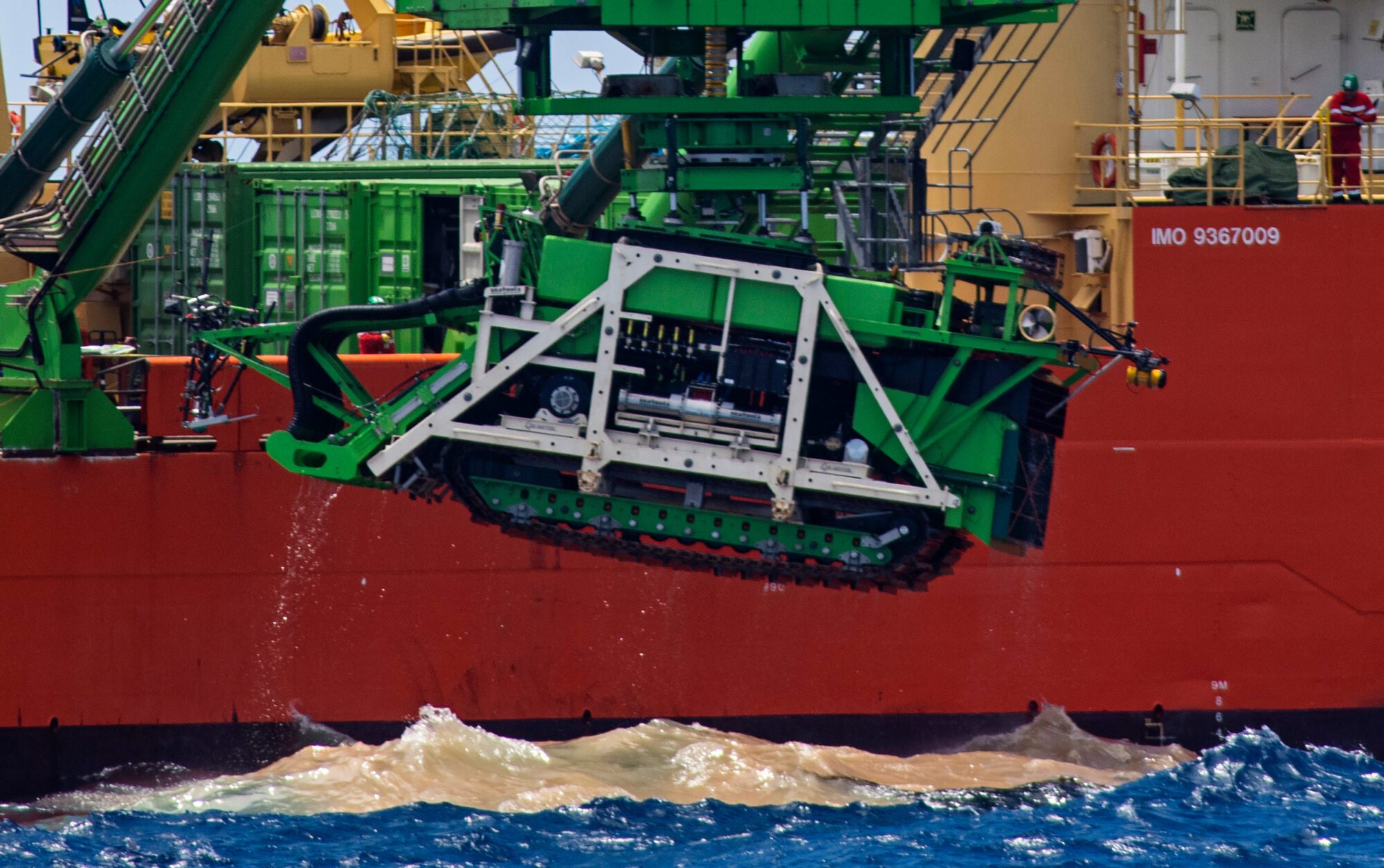 A side view of a large red ship filling the whole frame with a smaller vehicle that looks like a tank being lifted out of the ocean below. There is a large patch of yellow sediment visible in the dark blue water just below the machinery