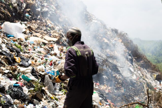 An African man in a boilersuit with hi-vis stripes and "care sanitation and suppliers ltd" written on the back of it stands looking out over a huge pile of smoking landfill rubbish towering over him. He is wearing a dirty face mask and a knitted cap, and thick gloves, holding crunched up Pepsi-branded plastic cups.