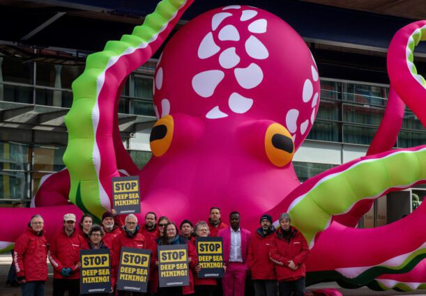 An inflatable pink octopus stands outside a tall hotel in London's canary wharf. A Greenpeace activist holds a banner reading 'STOP DEEP SEA MINING' in front.