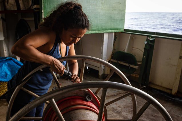 A woman attaches something to what looks like a hose on a very large metal reel