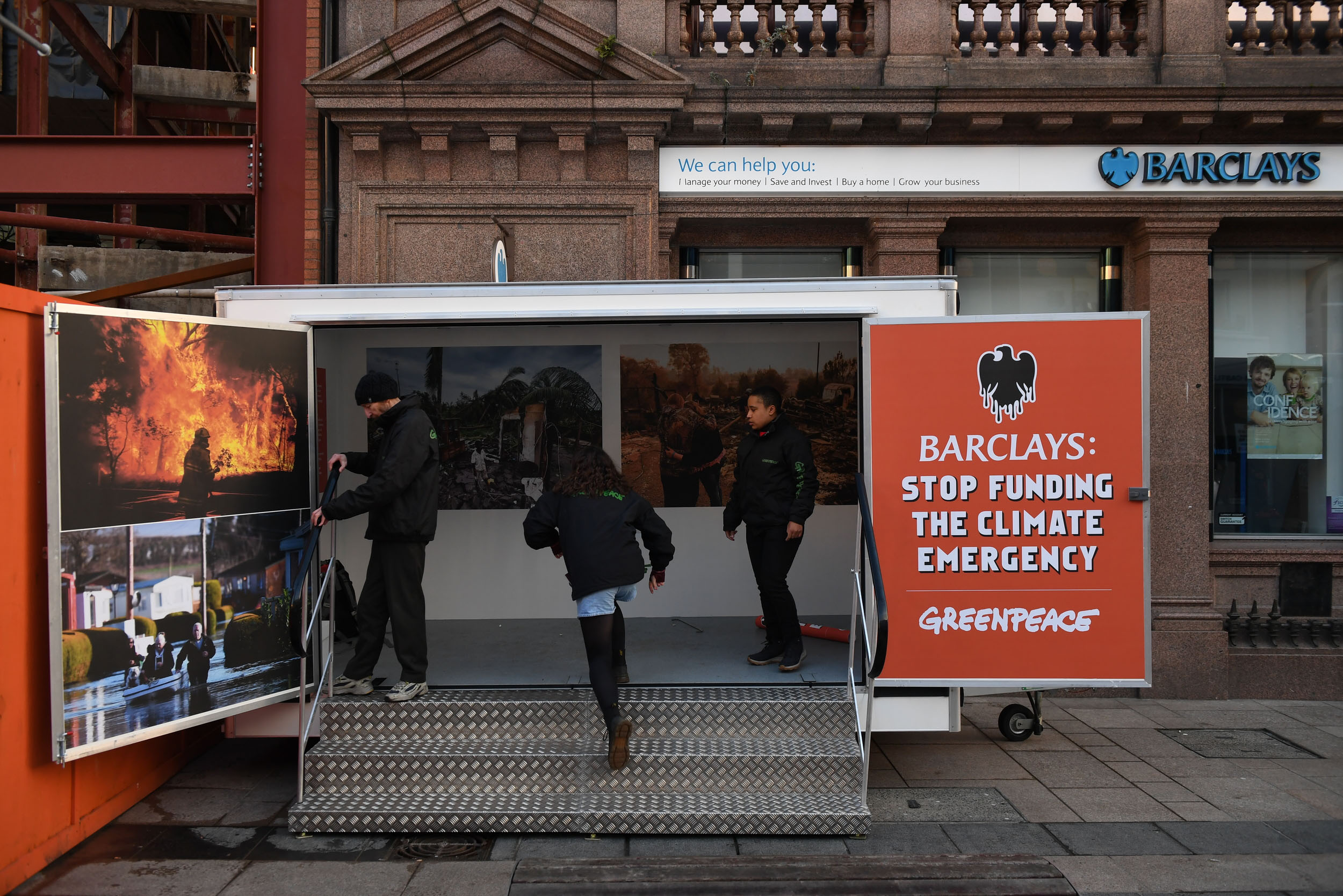 Members of the public look at photos of destruction caused by climate change, displayed in a temporary container placed in front of the entrance of a Barclays Bank branch
