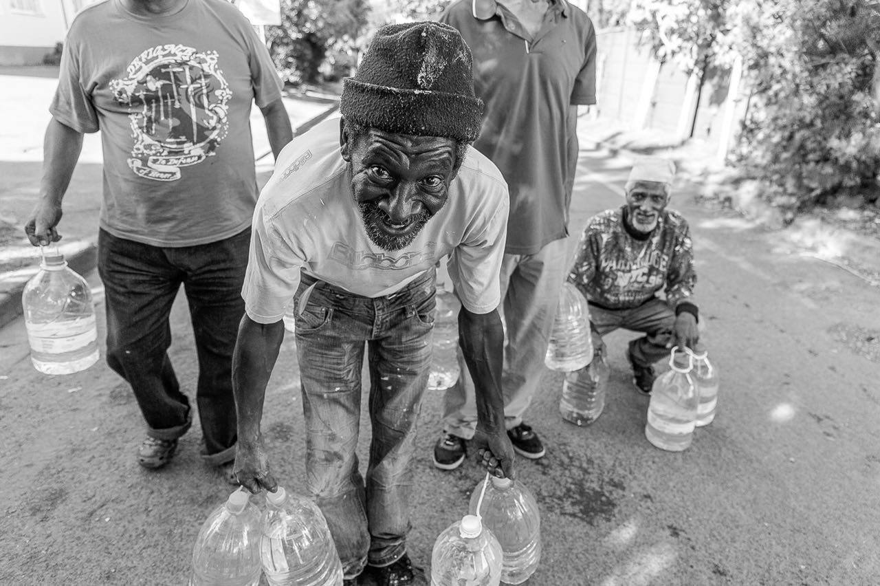 Four Black South African men, one in the foreground and one squatting so their faces show they are of older ages, holding five litre bottles of water to collect water. The men in front are holding at least four bottles, two in each hand, and smiling.