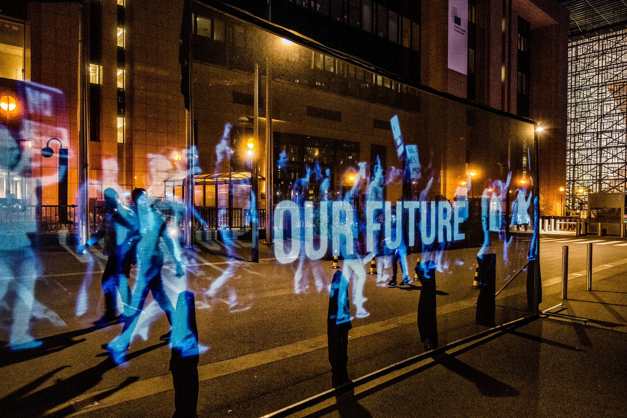 A holographic projection of people marching with a banner reading 'Our future', on a city street at night.