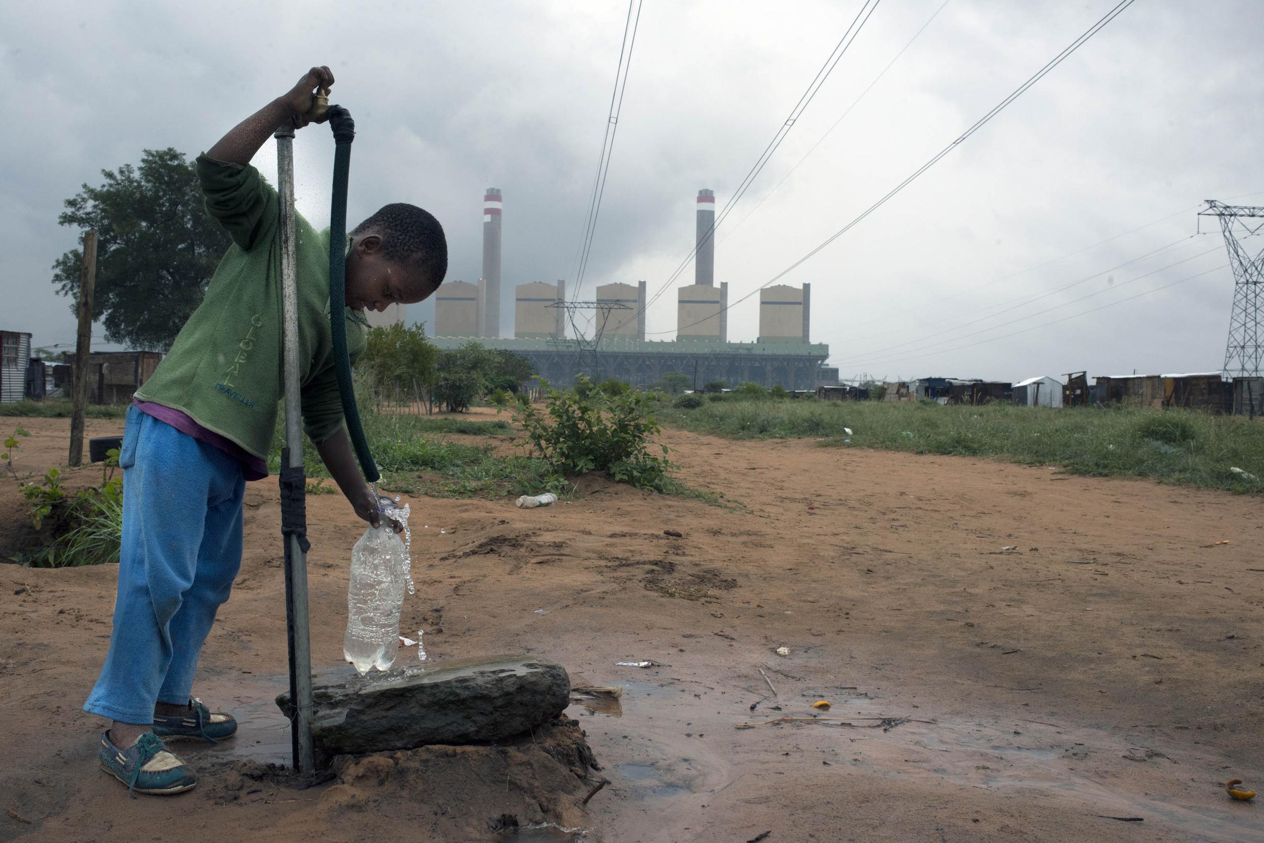 A small boy at a water tap in blue trousers and a green top. The red earth is wet around the tap and the sky is grey, in the background a power station with lines and pylons.