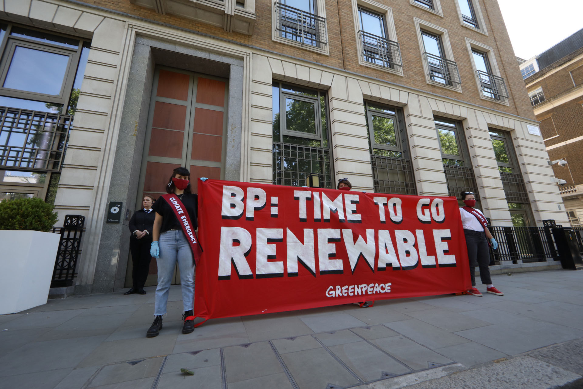 Two activists wearing masks and red sashes stand in front of BP's headquarters holding a large banner reading "BP: time to go renewable"