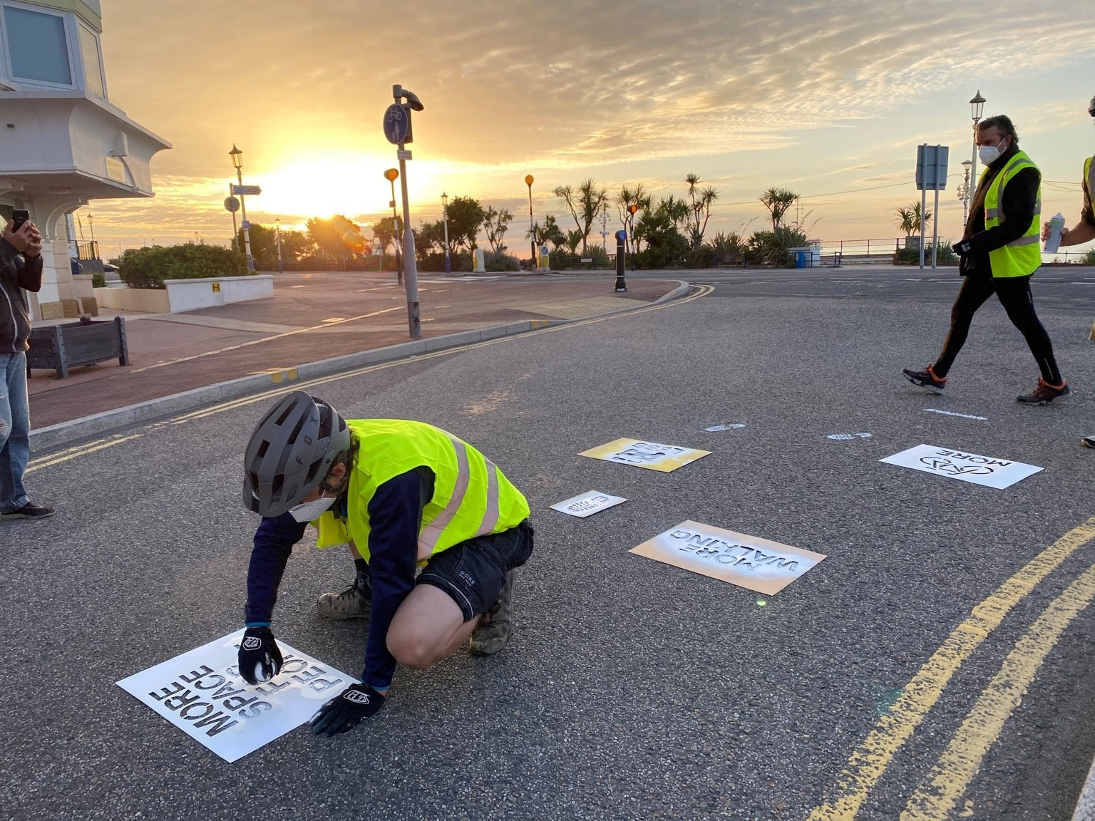 Greenpeace volunteers in fluorescent jackets apply stencils to a road surface, with a beautiful dawn sky in the background. 