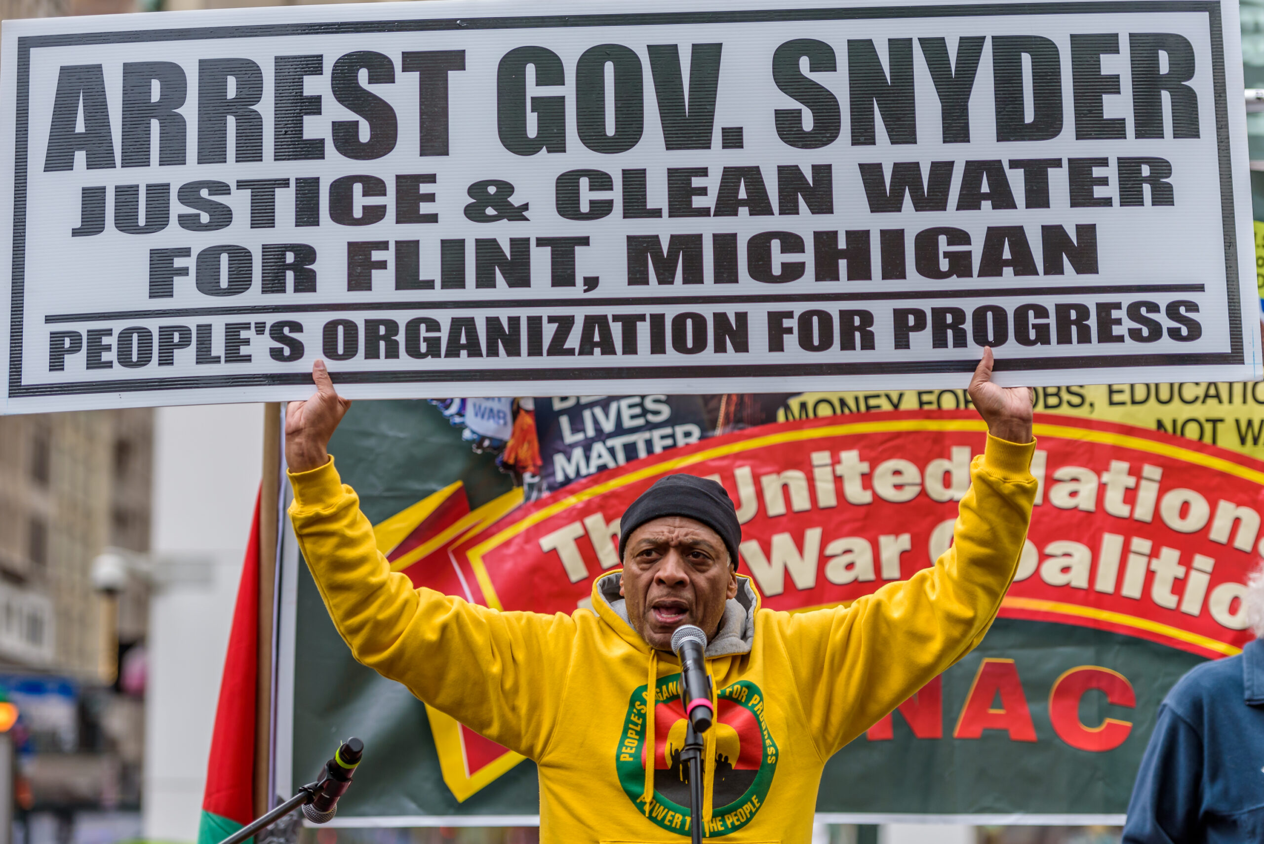 An older black man in a bright yello hoodie with an activist emblem on it holds up a sign saying 'Arrest Gov. Snyder. Justice & Clean Water for Flint, Michigan. People's Organisation for Progress'. He is in front of a banner on which the words 'united' and 'war' can be made out, and a sign saying 'black lives matter'