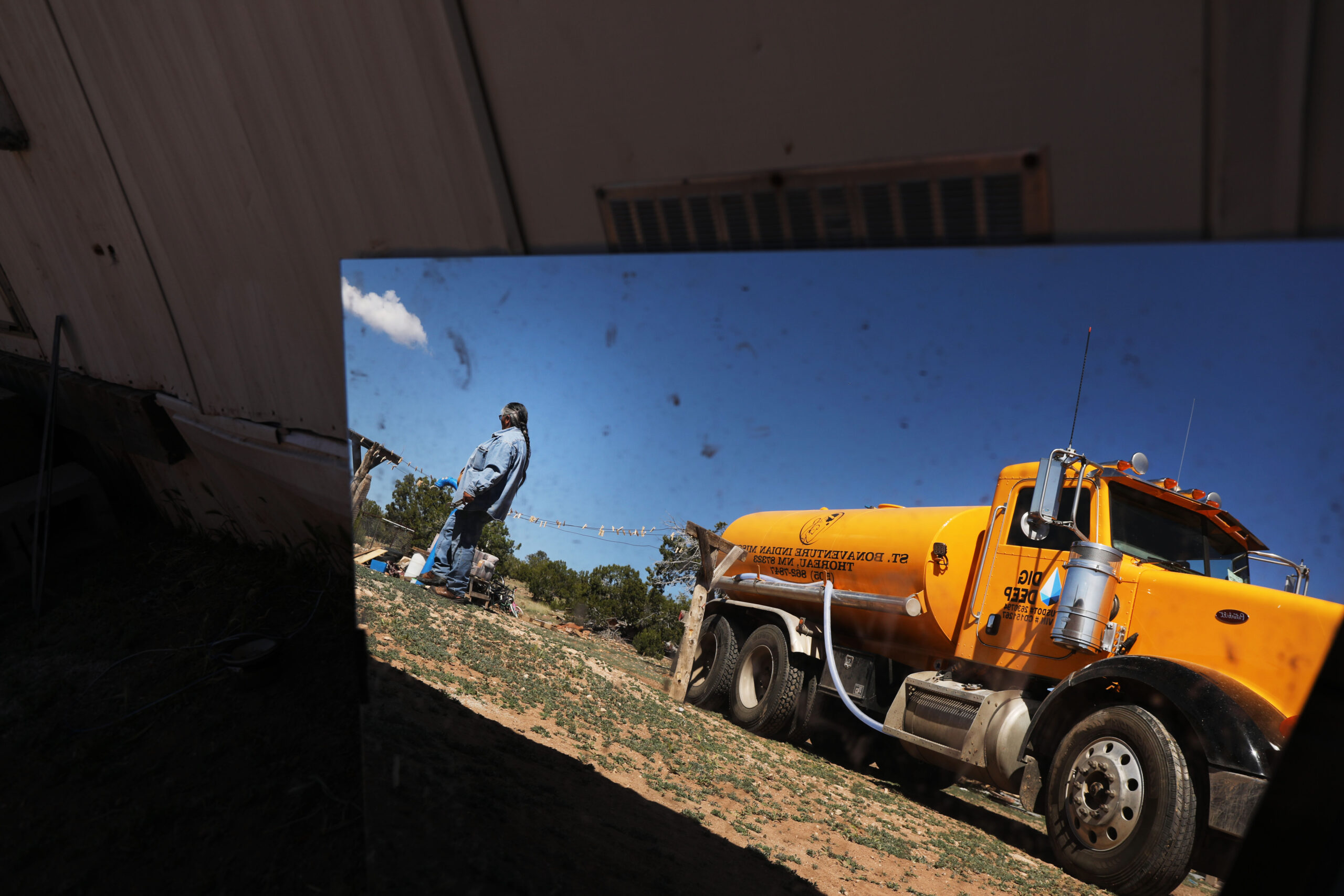 A large yello water truck with its operator reflected in a mirror, delivering water to a household on Navajo land.