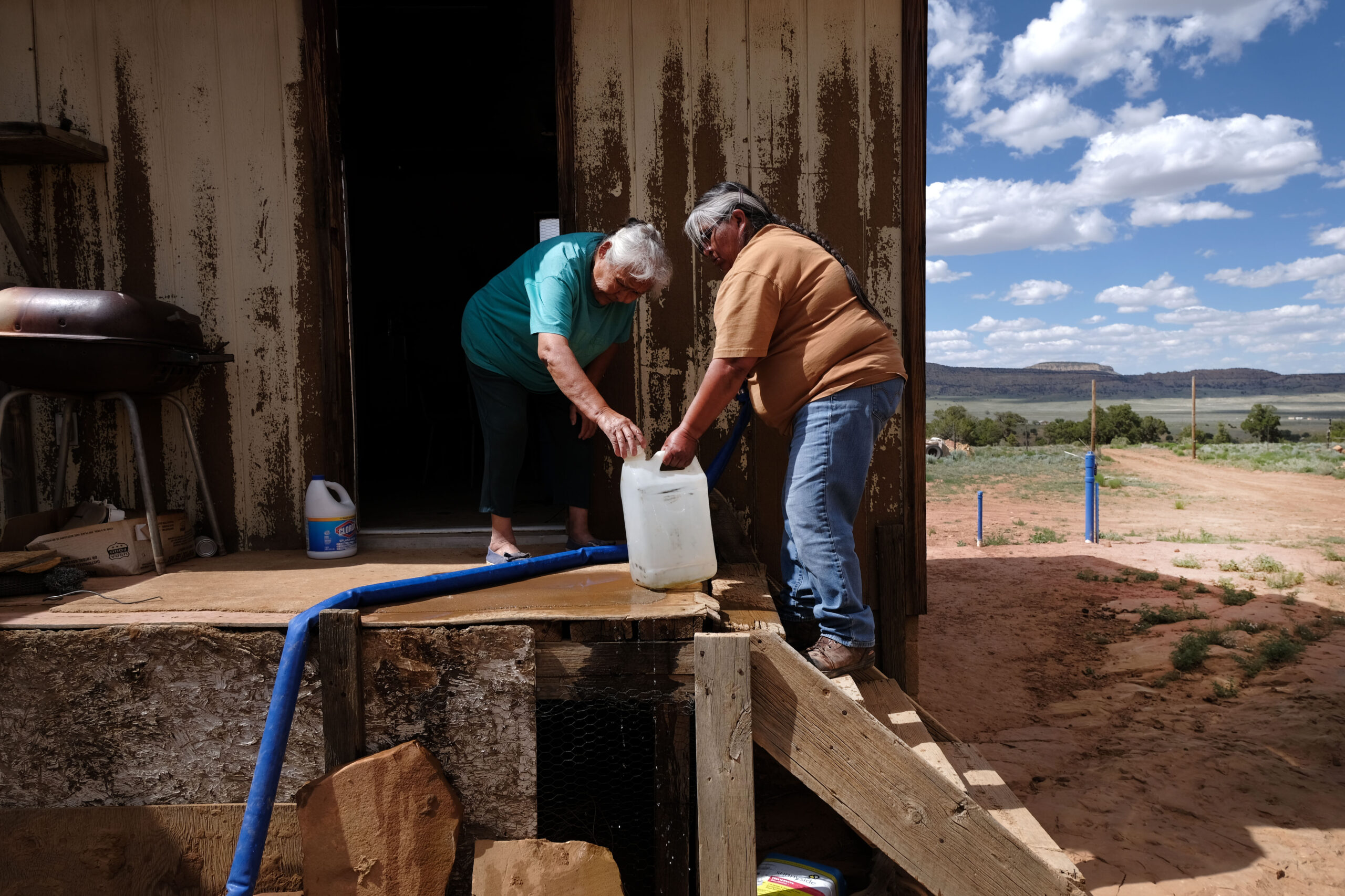 Two older Navajo women with a large plastic water bottle, outside a wooden clapboard house. There is a blue pipe running between them. The rural landscape stretches out into the distance.