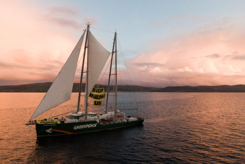 Aerial view of Greenpeace ship, the Rainbow Warrior, on the water at sunset. Between the sails is a large banner reading 