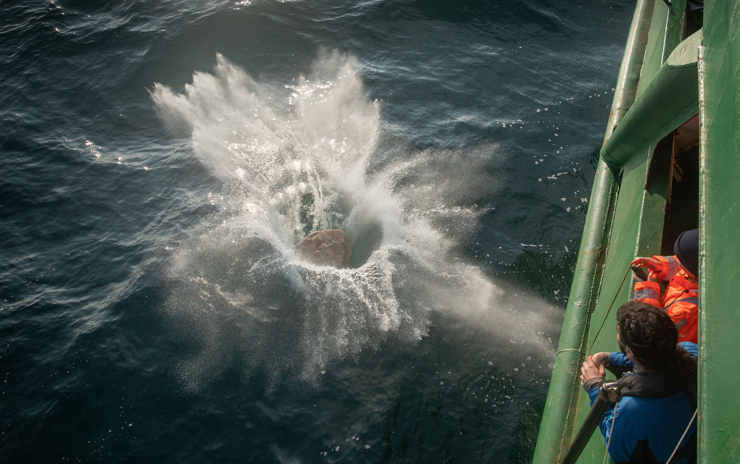 A boulder splashes into the water as crewmembers look on.