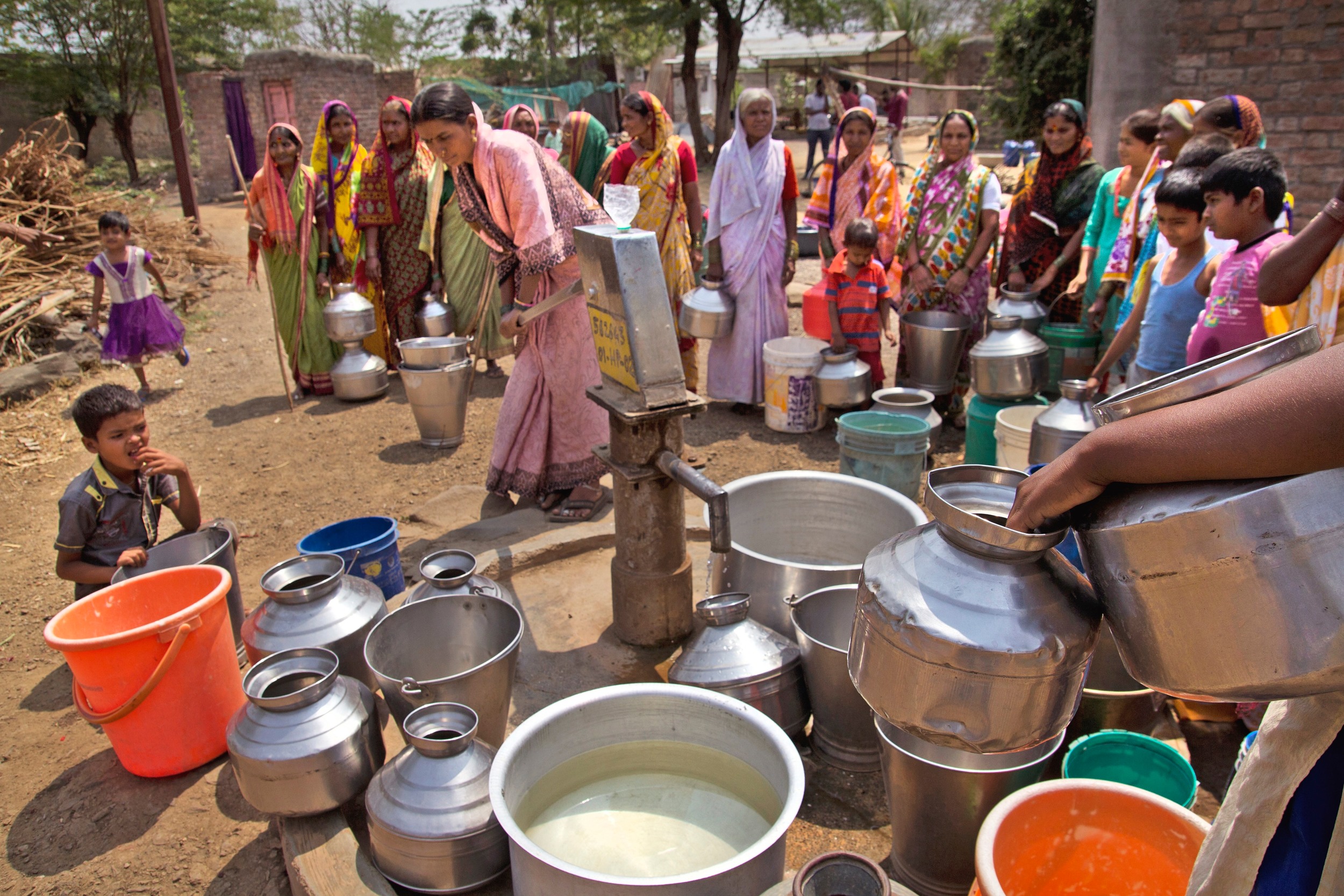 Around 17 Indian village women in colourful saris gather around a single tap in the centre of the frame. There are around 30 containers of water on the ground near the tap and in front of the women, made from metal, plastic and in different shapes and colours and sizes. There are two little children watching closely from the left hand side.