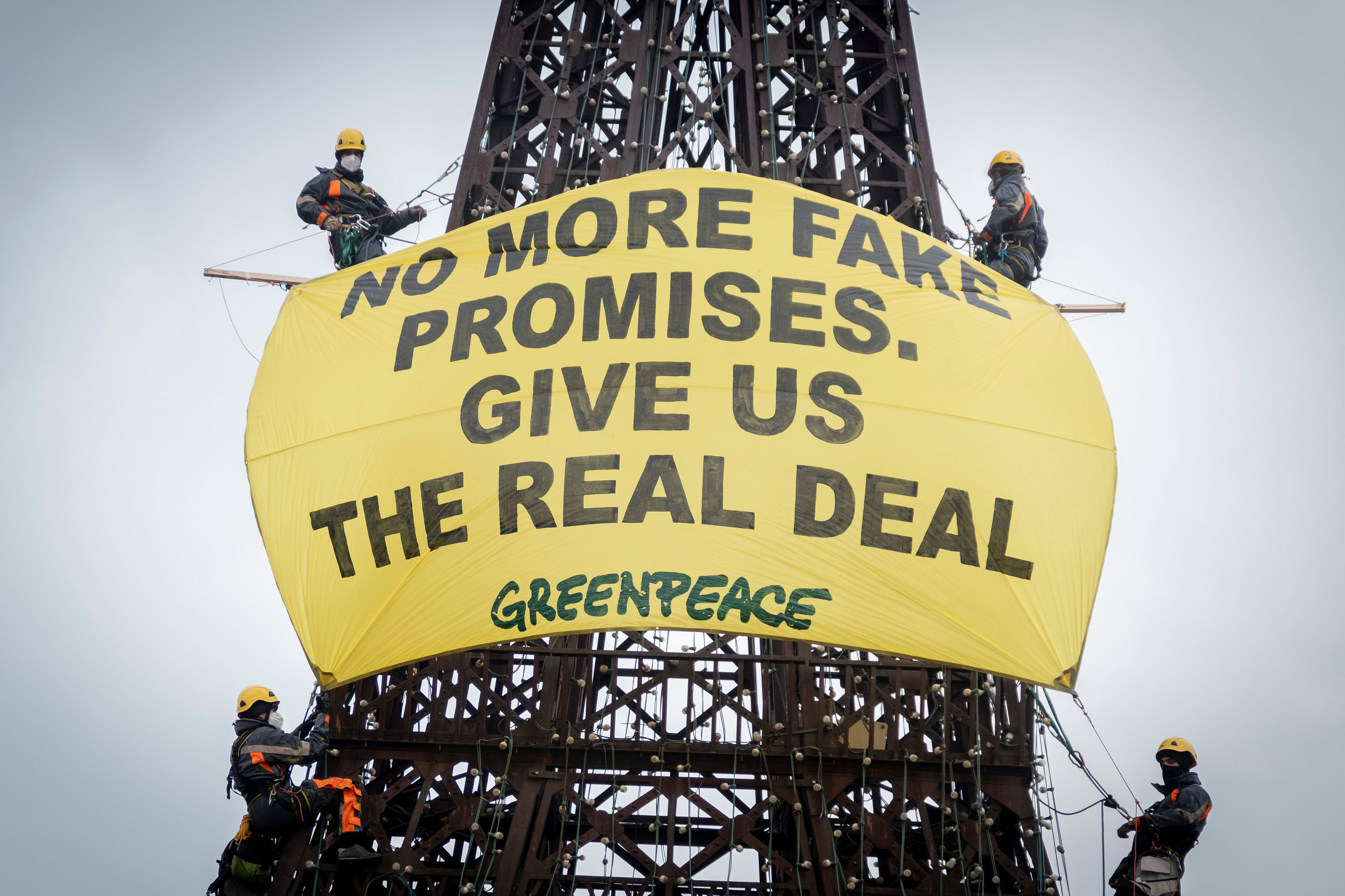 Greenpeace climbers in masks and helmets hang from a dark latticed metal structure, displaying a large yellow banner that reads "No more fake promises. Give us the real deal."