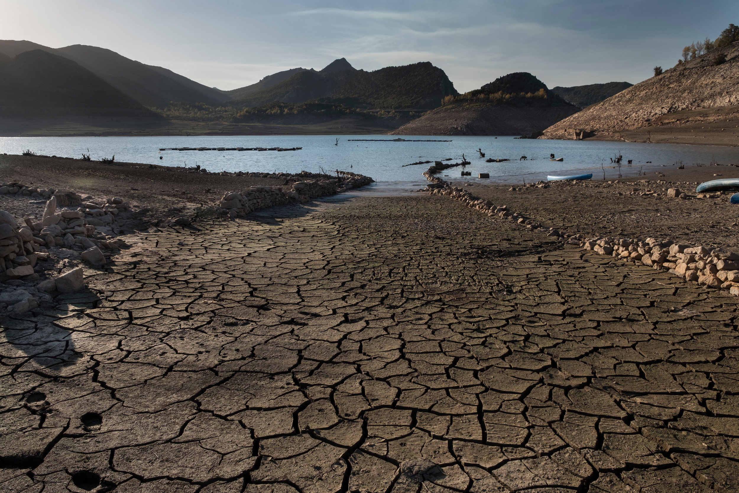 An extremely dried-out, brown landscape with cracked earth in the foreground, with a reservoir in the distance and mountains behind.